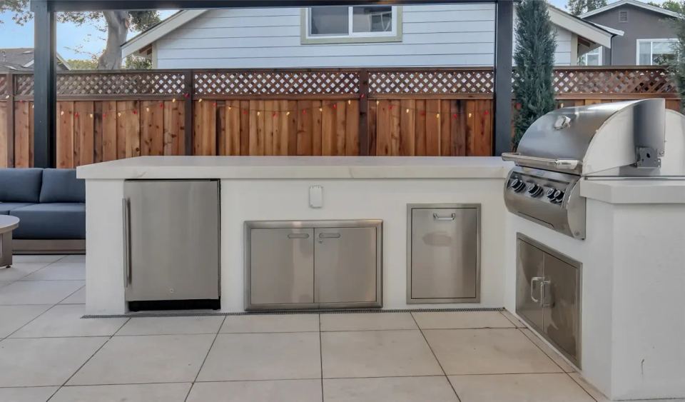 Enjoy your backyard remodel with an outdoor kitchen featuring stainless steel appliances, including a grill and refrigerator. It's all set under a patio surrounded by wooden fencing, perfectly complementing the light gray house in the background.