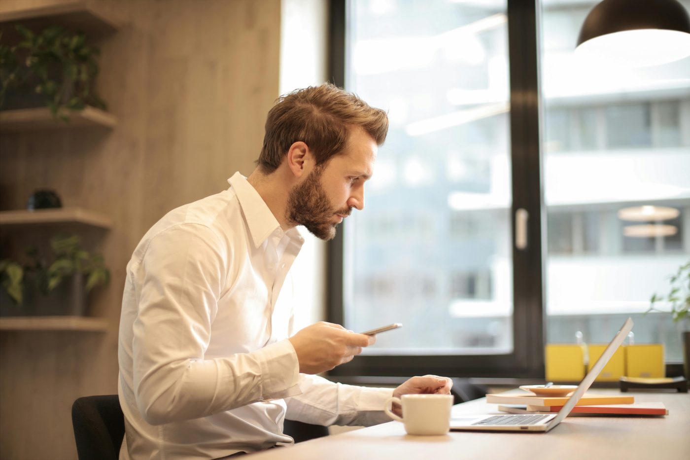 A man is looking at his laptop while holding a smartphone.