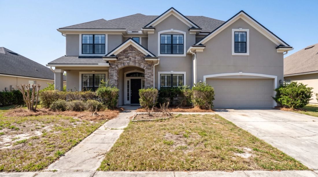 A two-storey Florida house with a patchy lawn, overgrown shrubs and bare beds