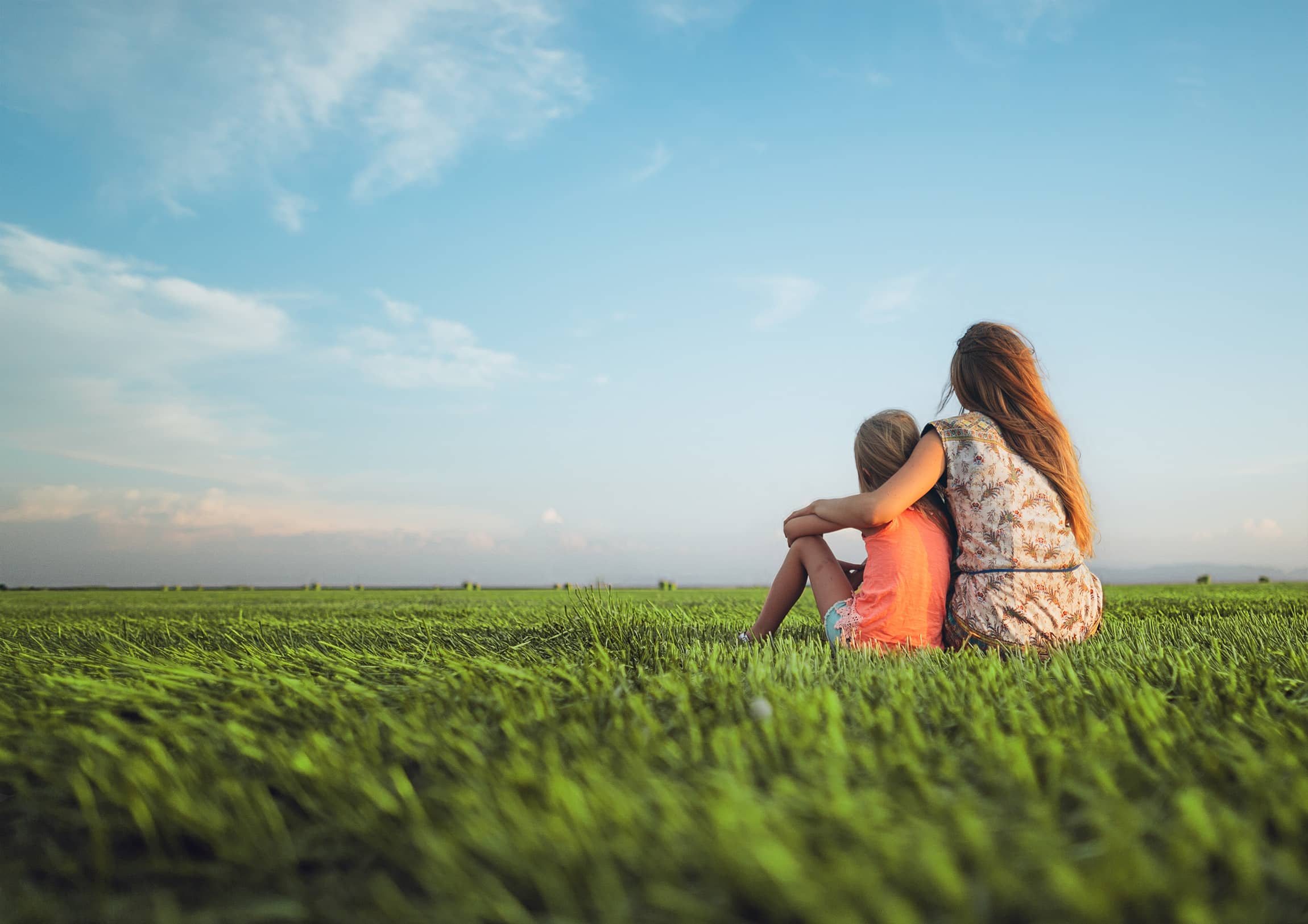 A woman sits in lush grass with her child as they look out at the blue sky and fluffy clouds on the horizon