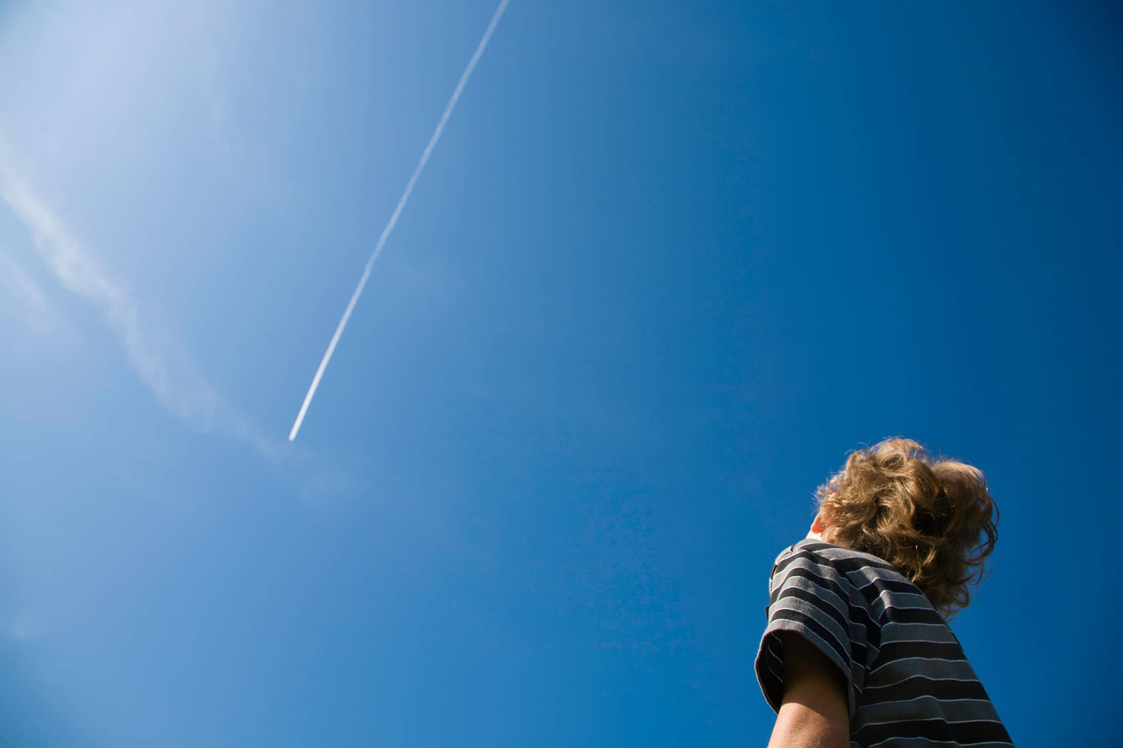 A young child watches a plane create a contrail against a clear, blue sky.