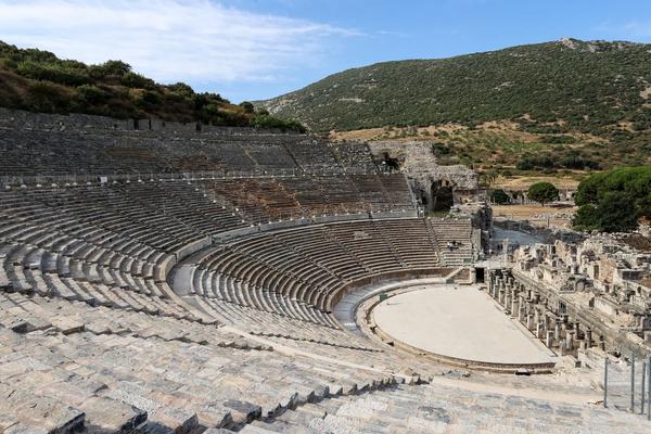 A panoramic view of the ancient Great Theatre of Ephesus, featuring its massive semicircular stone seating built into a steep hillside. The flat, semi-circular orchestra area at the base sits before the ruins of a colonnaded stage building, with rugged green mountains and a blue sky in the background.