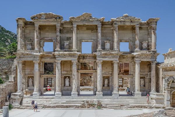 The two-story marble facade of the ancient Library of Celsus in Ephesus. The structure features ornate Corinthian columns, decorative triangular and curved pediments, and statues nestled in niches. A few tourists are scattered across the stone courtyard and within the entranceways under a clear, bright blue sky.