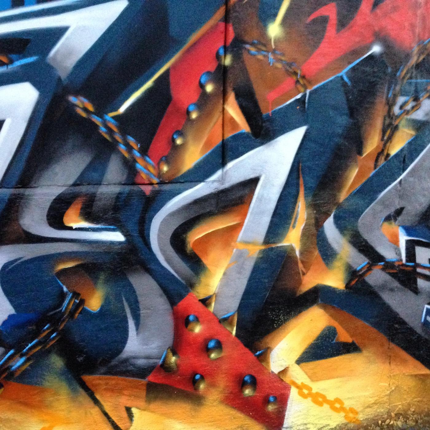 Detalj med graffitibokstäver och kedjor som speglar eldsken på Spawn-väggen. Detail of graffiti letters with chains reflecting firelight on the Spawn tribute wall.