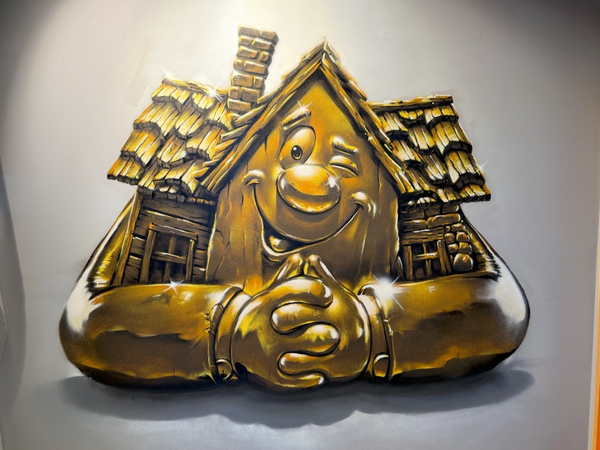 Gyllene 3D-maskot i form av huset Lebe, målad på en vit vägg vid entrén till XL Bygg Lebe Byggvaror i Tyresö, fotorealistisk målning av Spraya. Golden 3D house mascot Lebe painted on a white wall at the XL Bygg Lebe Byggvaror entrance in Tyresö, a photorealistic mural by Spraya.