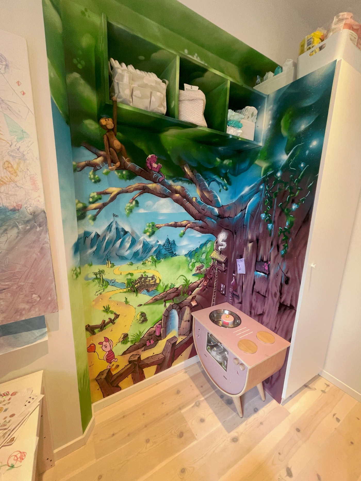 Hörn i ett barnrum i Stockholm målat som en magisk skog med ett stort träd, berg och en äng, inspirerat av Nalle Puh. Kids' room corner in Stockholm painted as a magic forest with a big tree, mountains and a meadow, inspired by Winnie-the-Pooh.