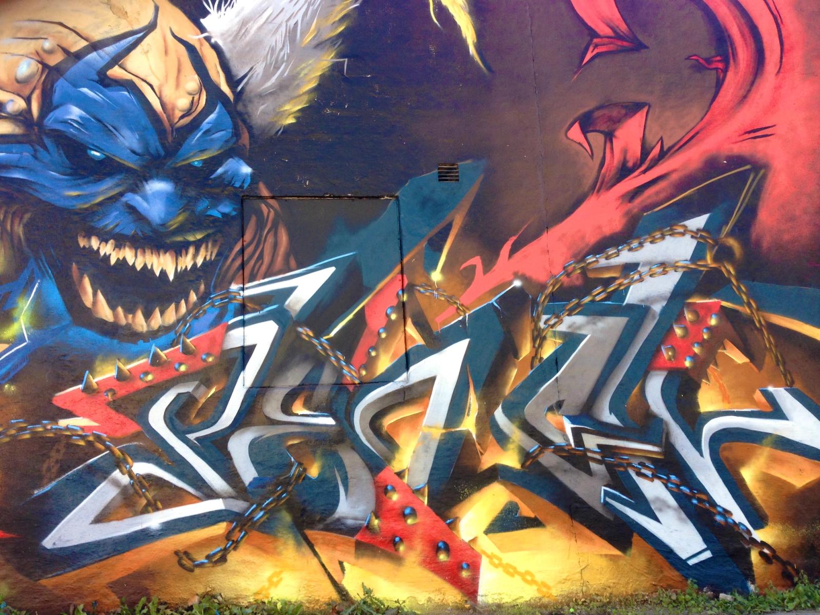 Graffitivägg i Snösätra med en flinande Violator ur Spawn-serien, brinnande graffitibokstäver och kedjor, målad på Springbeast 2014. Graffiti wall in Snösätra with a grinning Violator from the Spawn comics, burning graffiti letters and chains, painted at Springbeast 2014.