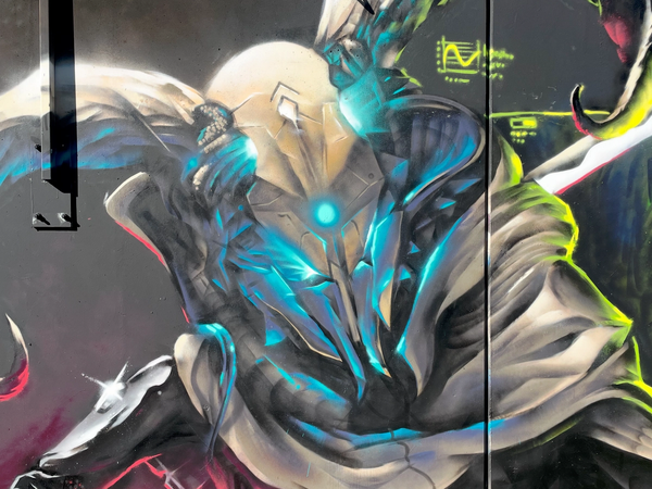 Sci-fi-graffitimålning i Visby av Spraya: en mantelklädd utomjording med lysande blått ansikte och spjut, ur The Raukonium Saga. Sci-fi graffiti mural in Visby by Spraya: a cloaked alien council member with a glowing blue face and a spear, from The Raukonium Saga.
