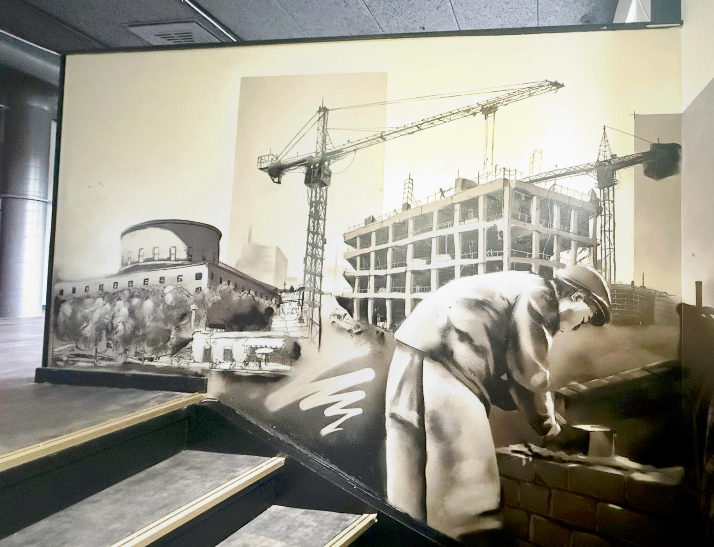 En murare i arbete framför lyftkranar och ett hus under uppbyggnad, ur Construction homage i Tyresö. A bricklayer at work in front of cranes and a building under construction, from the Construction homage mural in Tyresö.