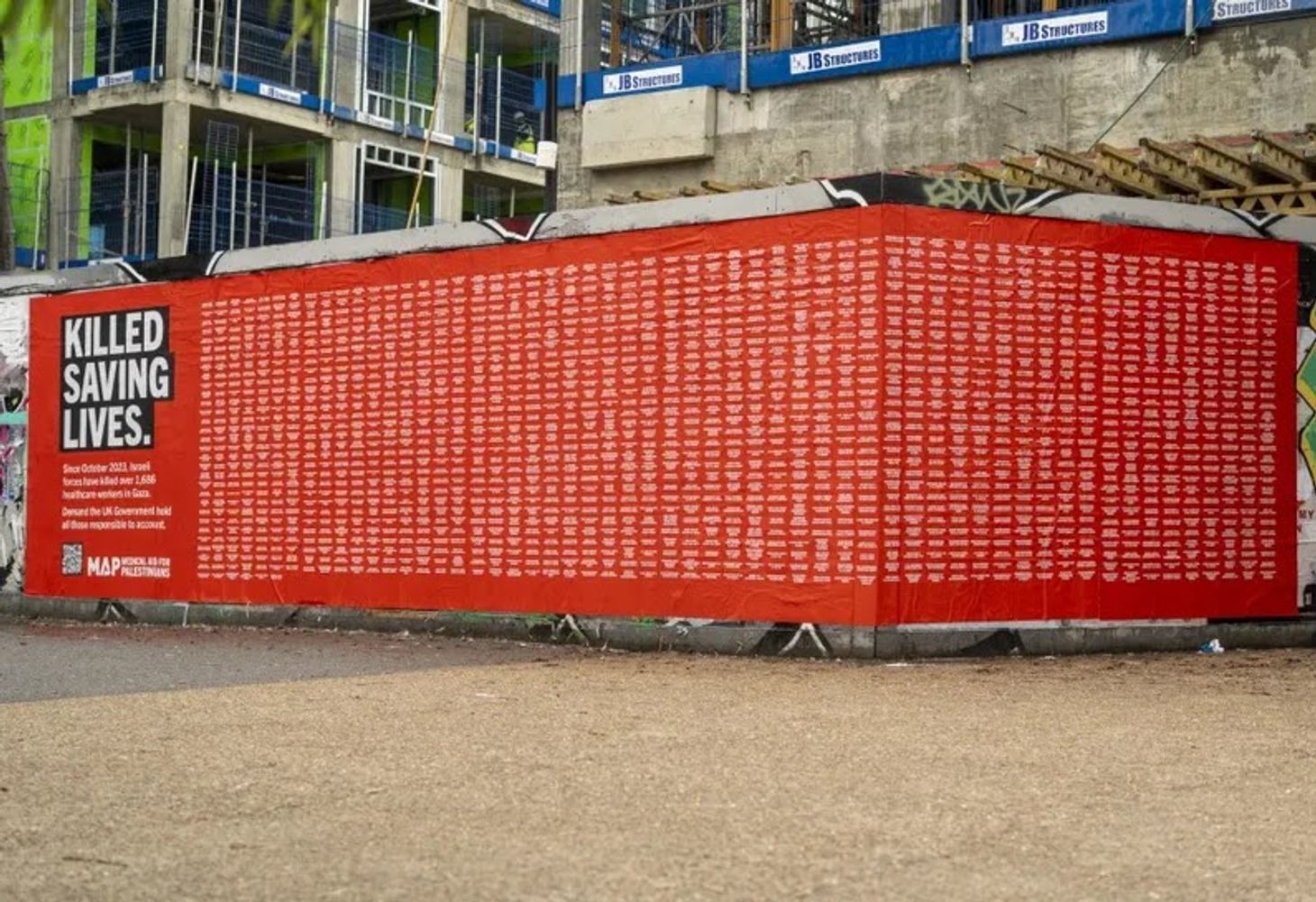 Red building site hoarding covered with the names of Palestinian health workers killed in Gaze, with bold text reading “Killed Saving Lives.”
