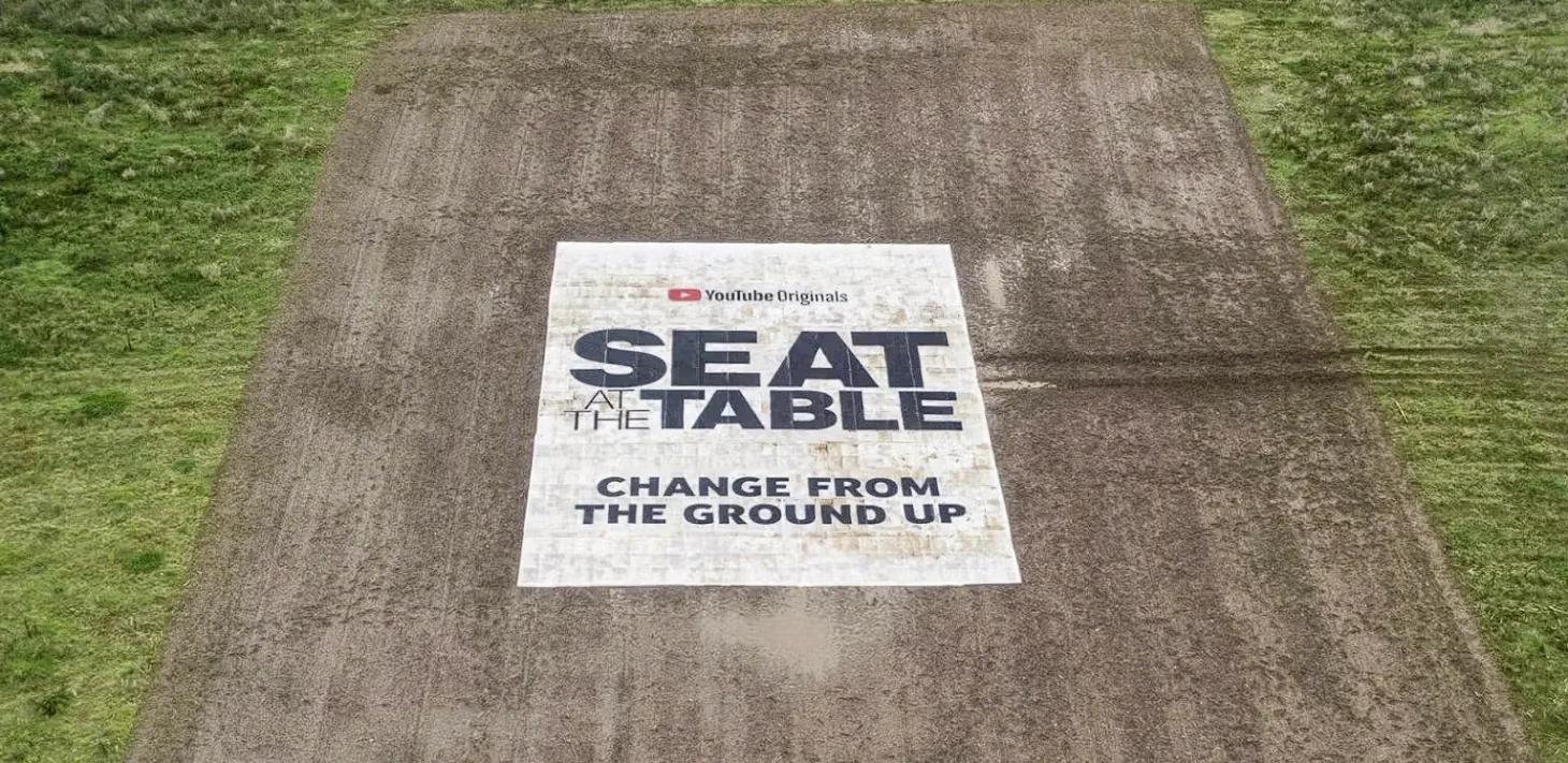Aerial view of a plowed field with a white box with text in the centre reading “Seat at the Table, Change from the ground up”.