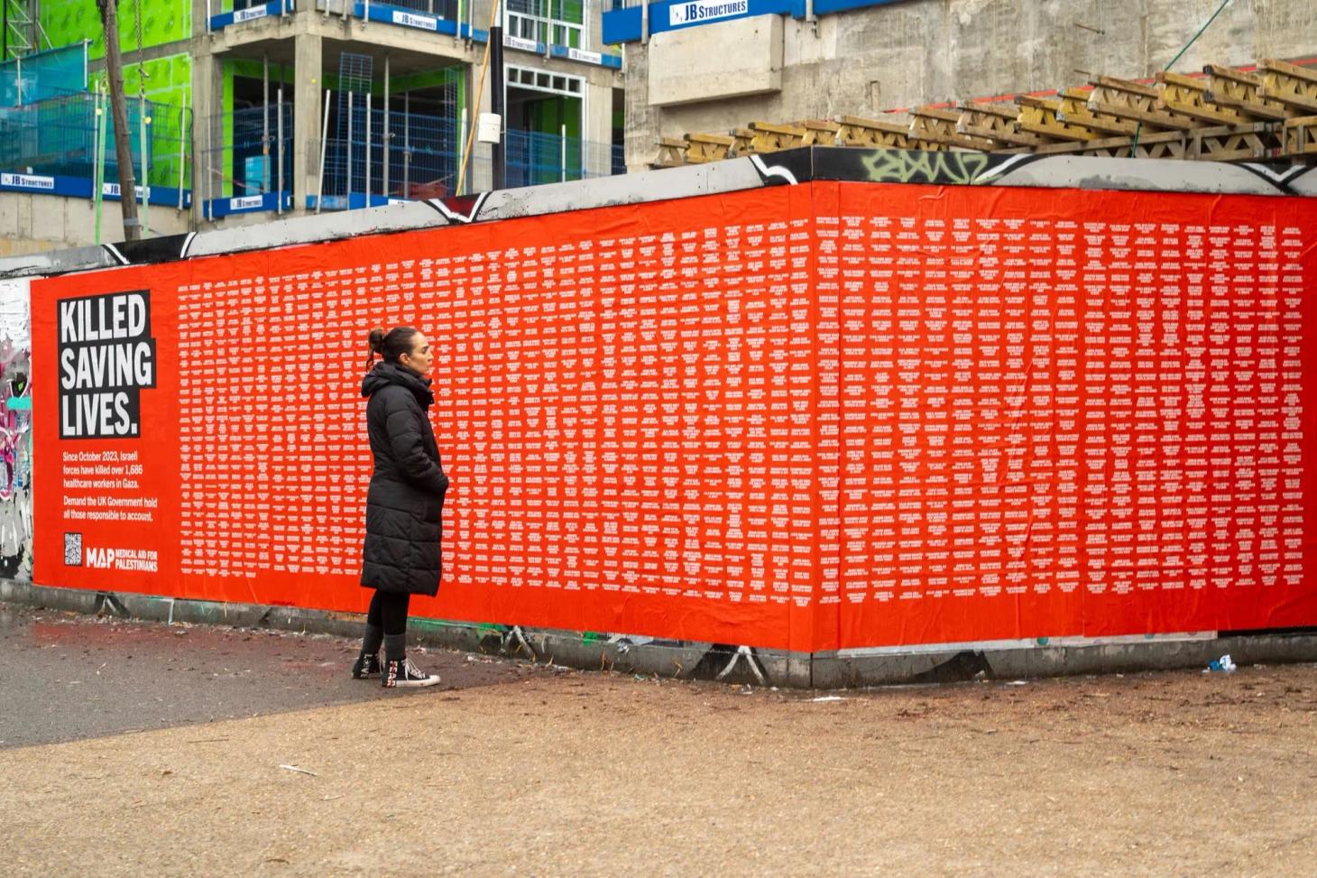 Woman pausing at MAP's red Killed Saving Lives wall naming 1,686 Gaza health workers