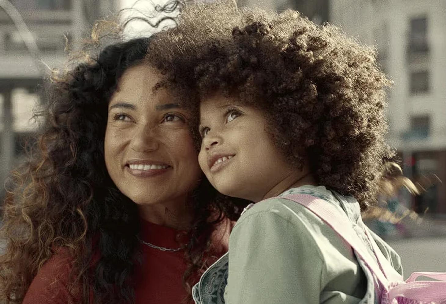 A woman and a child with curly hair, embracing and smiling while looking upwards. The background is an urban setting.