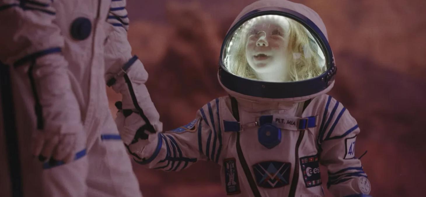 A young blonde girl in a lit-up space suit gazes upward with wonder, holding hands with another suited figure against a reddish backdrop on another world.
