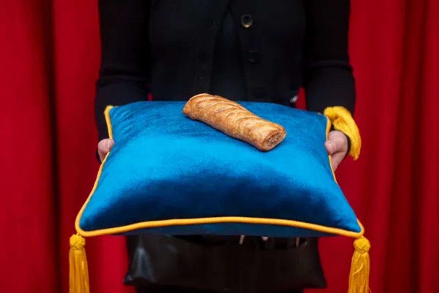 A wax-work Gregg’s sausage roll presented on a royal blue cushion