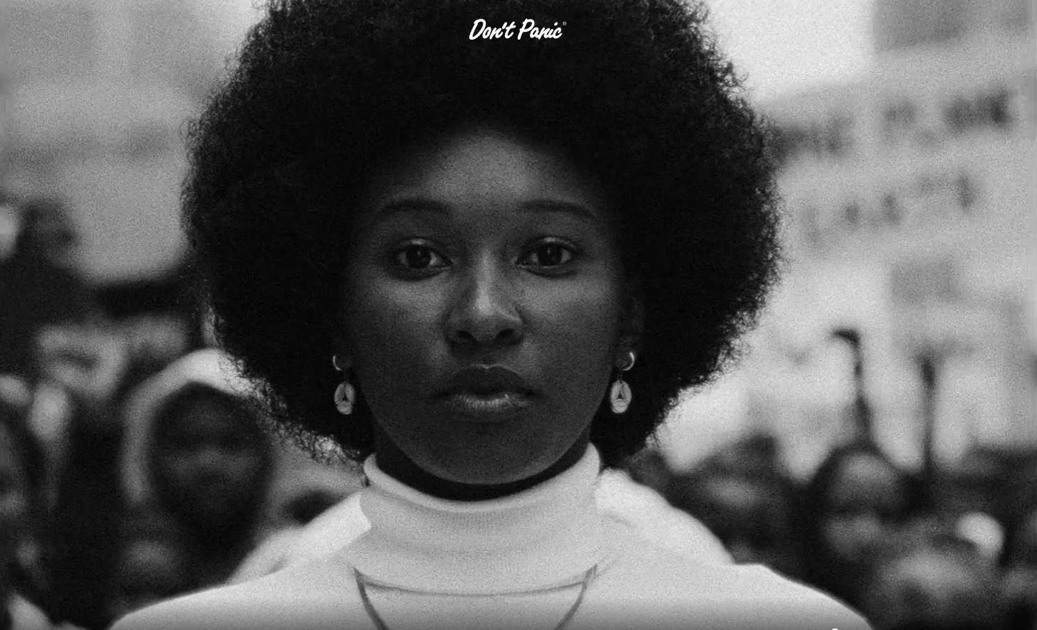 A black woman in a white polo neck stands in at the front of a protesting crowd, looking defiantly at the viewer
