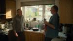 Two middle-aged men talking in a bright kitchen, one leaning on the counter holding a dish towel.