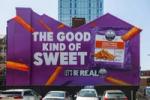 Front-on view of a large purple Strong Roots mural in Manchester, created by Don’t Panic London, reading “The Good Kind of Sweet” beside an oversized pack of crispy sweet potato fries.