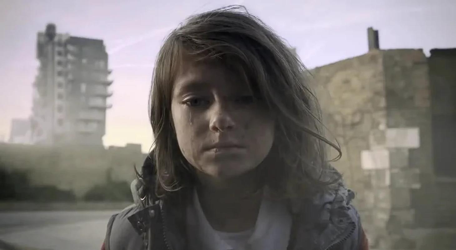 A young girl with tousled hair and a tear-streaked face stands in a desolate urban area.