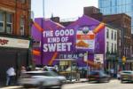 Wide street view of Don’t Panic London’s Strong Roots mural in Manchester, featuring “The Good Kind of Sweet,” an oversized product pack and orange fries, with pedestrians and blurred traffic below.
