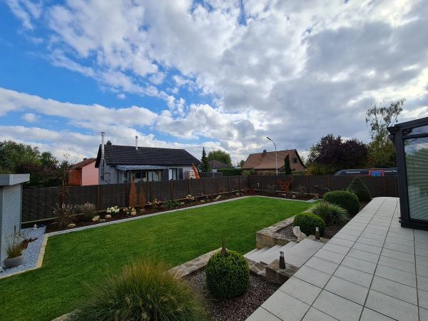 Gestalteter Garten mit braunem Flechtzaun, Rasen, Beeten und Terrasse aus hellen Platten