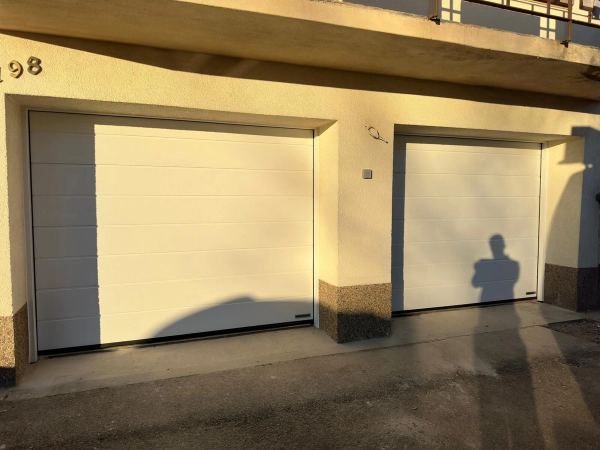 Zwei weiße Sektionaltore unter einem Balkon, cremefarbene Fassade in der Abendsonne