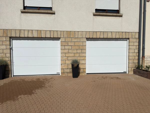 Zwei weiße Sektionaltore nebeneinander in einem Natursteinsockel, gepflasterter Hof