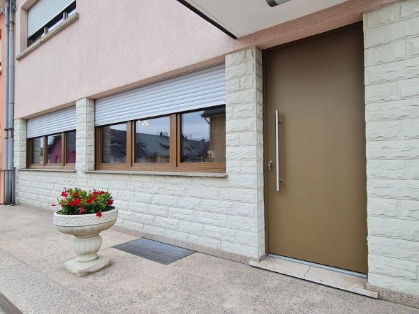 Glatte bronzefarbene Haustür mit Edelstahl-Stangengriff, weiße Steinfassade und Fenster mit Rollläden