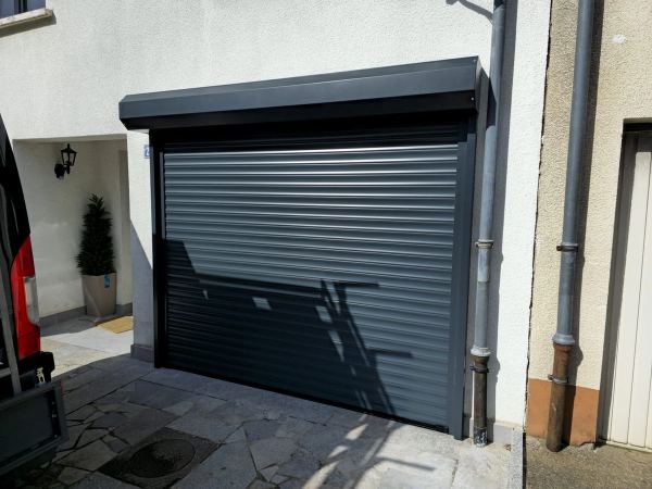 Anthrazitfarbenes Rolltor mit sichtbarem Kasten an weißer Fassade über gepflastertem Hof