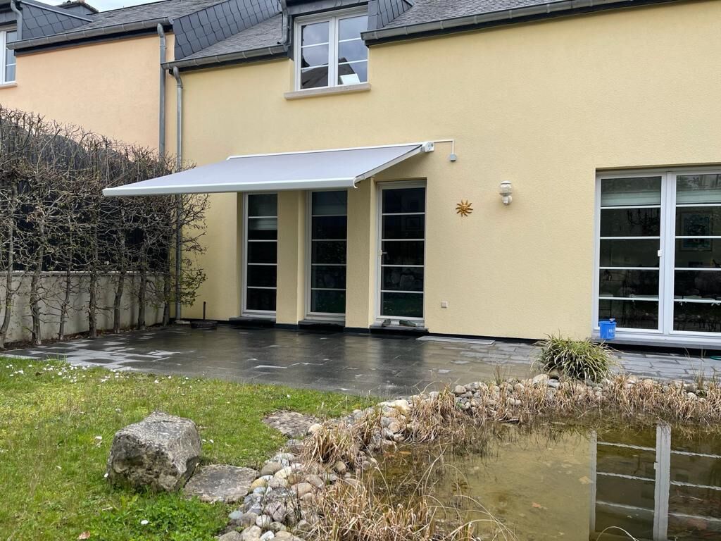 Hellgraue Markise über Terrassentüren eines gelben Hauses mit Gartenteich