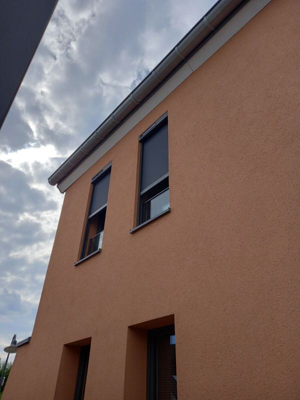 Graue Senkrechtmarkisen halb heruntergelassen an zwei Fenstern, orange Fassade