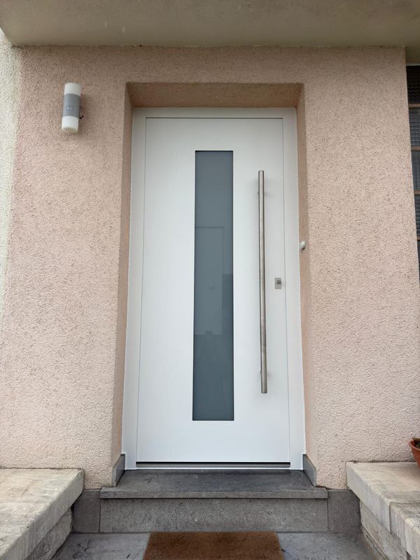 Weiße Haustür mit satiniertem Glasstreifen und langem Edelstahlgriff, rosa Putzfassade und Steinstufe