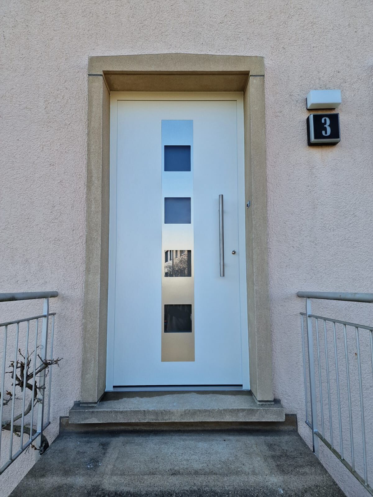 Weiße Haustür mit Edelstahlstreifen und vier quadratischen Glasfeldern, Steinrahmen in rosa Fassade