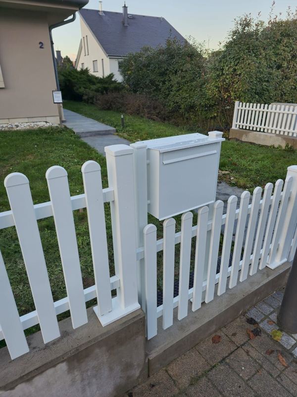 Weißer Lattenzaun auf einer Mauer mit integriertem weißem Briefkasten vor einem Garten