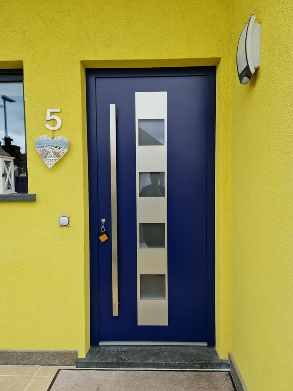Dunkelblaue Haustür mit Edelstahlstreifen, vier quadratischen Glasfeldern und langem Griff, gelbe Fassade