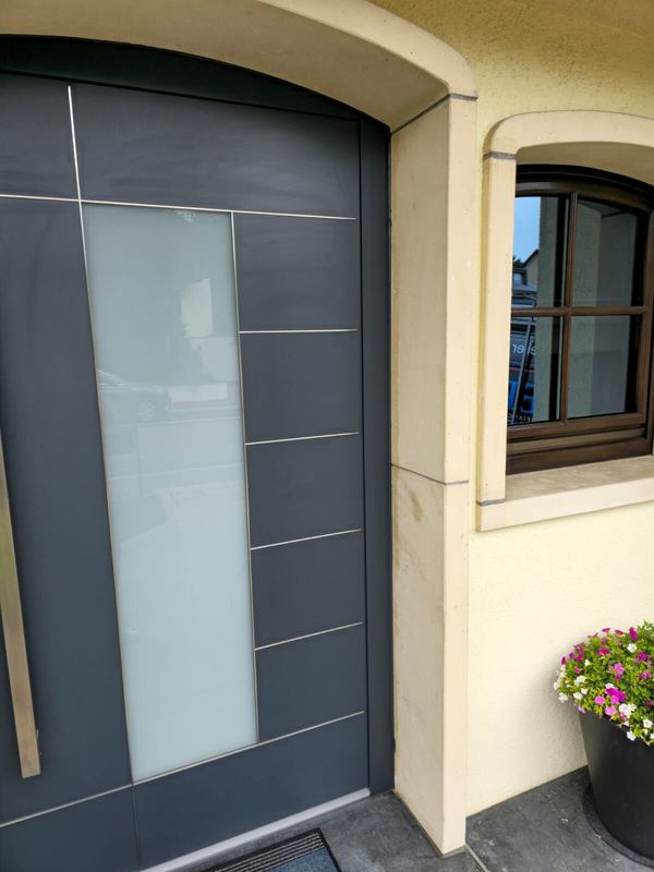 Detail einer anthrazitfarbenen Rundbogen-Haustür mit Milchglas und Edelstahllinien in cremefarbenem Steingewände