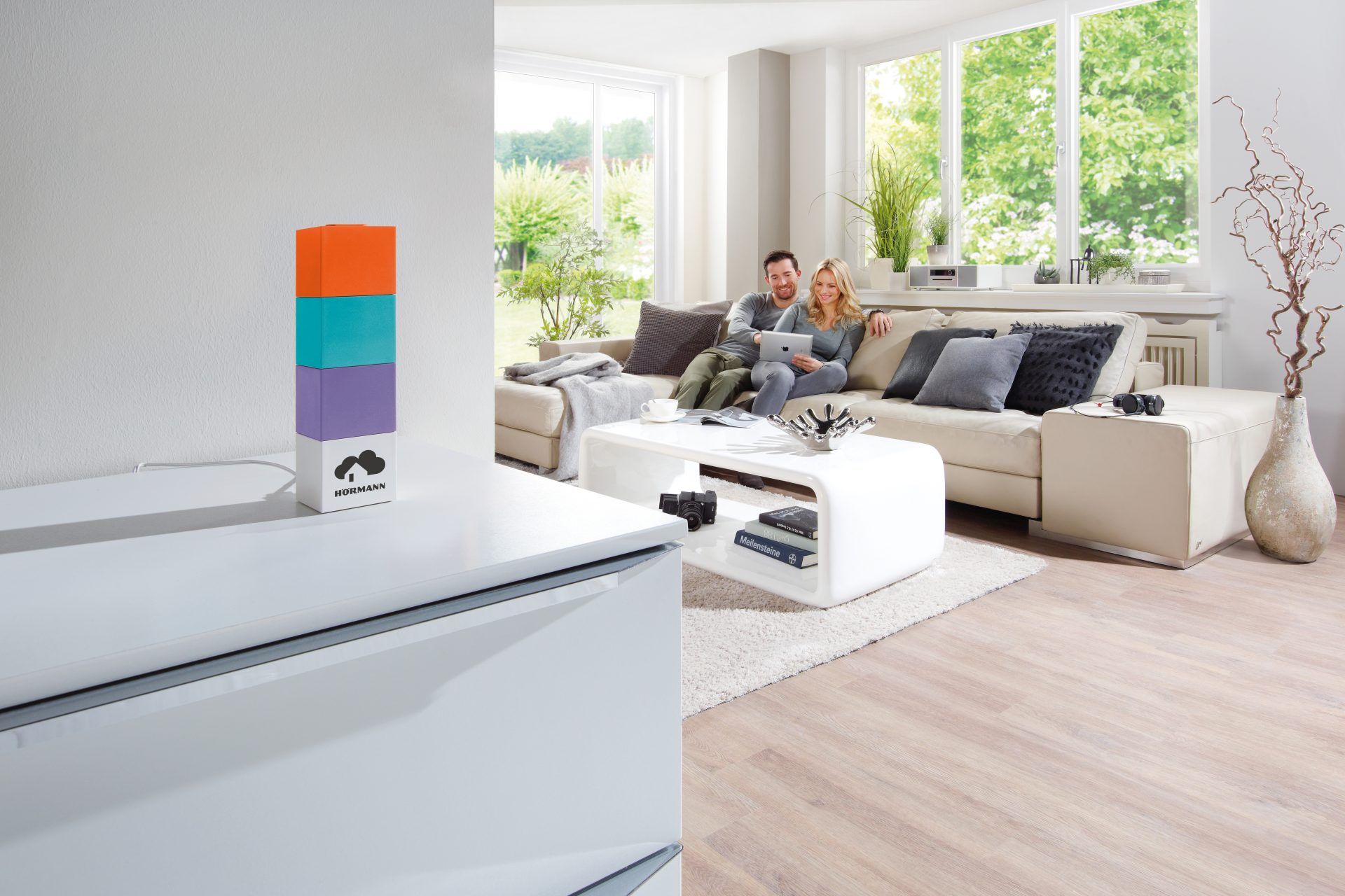 Smart-Home-Zentrale Homee mit farbigen Würfeln in einem hellen Wohnzimmer, ein Paar nutzt ein Tablet auf dem Sofa
