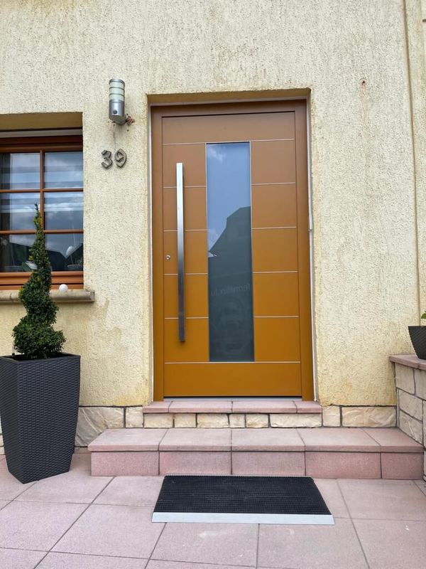 Ockerfarbene Haustür mit waagerechten Rillen, senkrechtem Glasstreifen und Edelstahlgriff über rosa Steinstufen