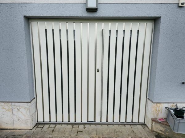 Weißes Flügel-Garagentor mit senkrechten, offenen Lamellen und mittigem Edelstahlgriff an blaugrauer Putzfassade