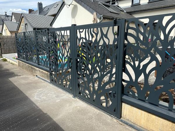 Anthrazitfarbene Gartentür und Zaun mit Blattmotiv-Ausschnitten auf einer Mauer am Gehweg