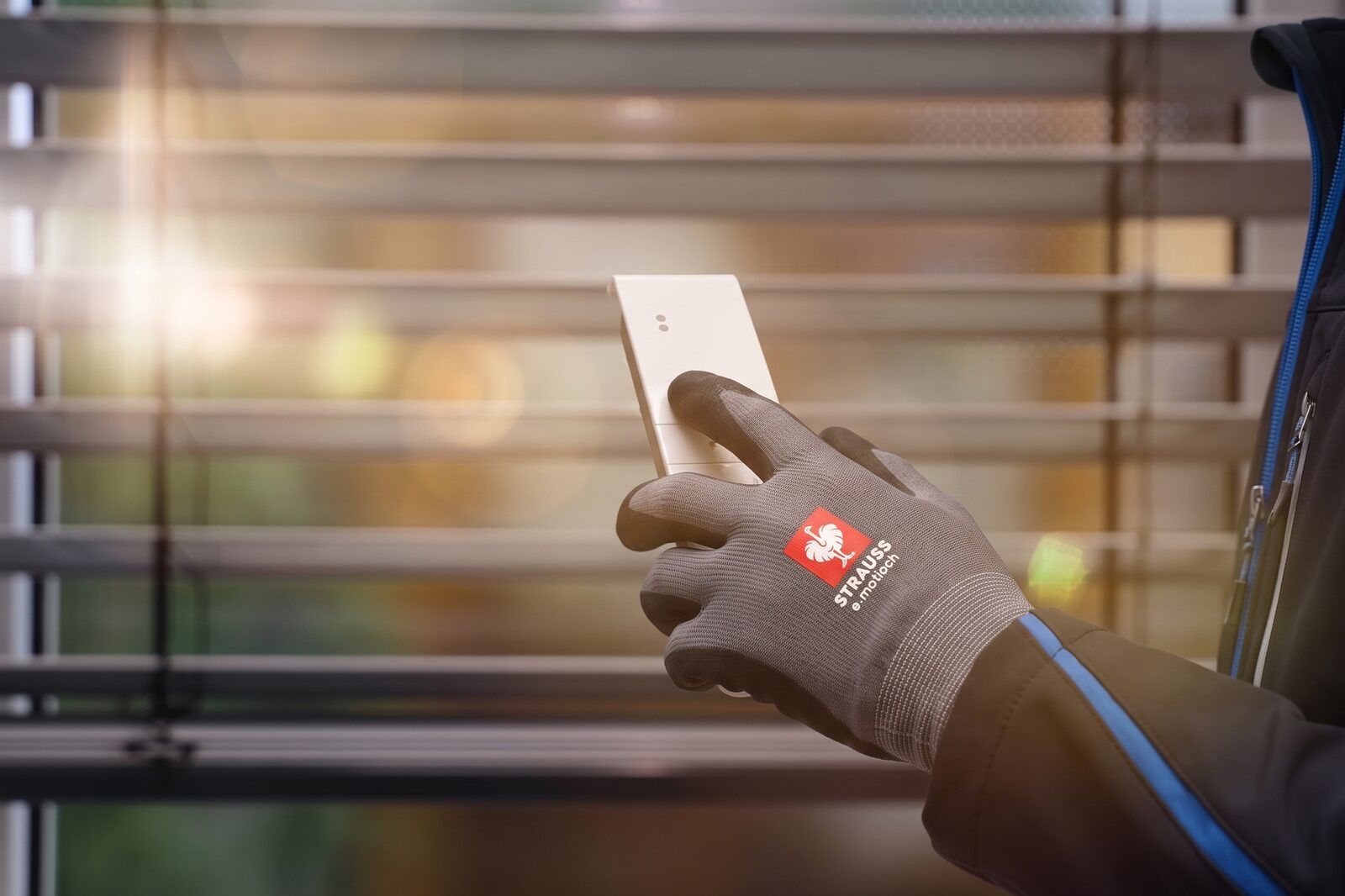 A technician’s gloved hand holding a wall transmitter in front of an external venetian blind