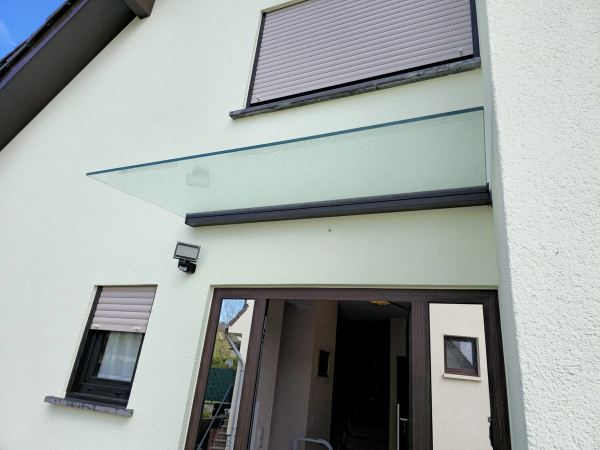 Vordach aus satiniertem Glas über einem verglasten Eingang mit braunem Rahmen, Putzhaus mit Rollläden