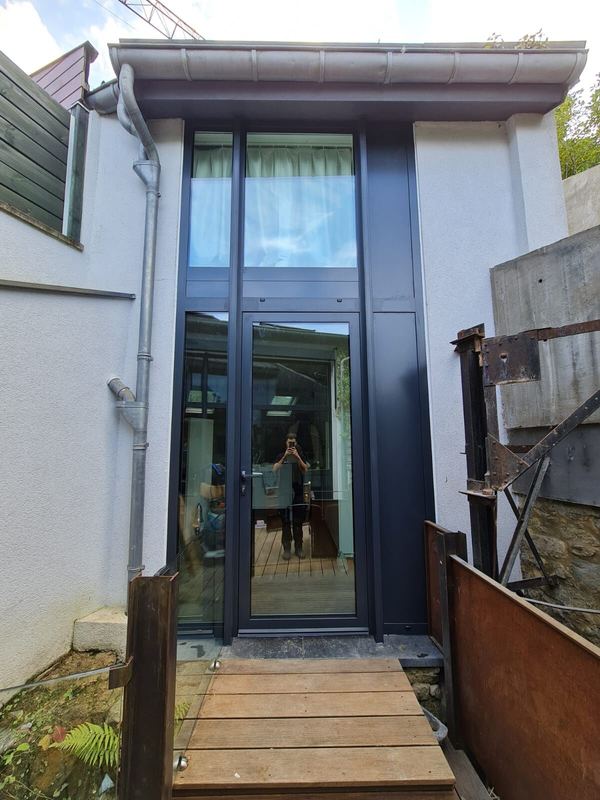 Zweigeschossige anthrazitfarbene Glasfront mit Eingangstür aus Glas, Zugang über Holzsteg
