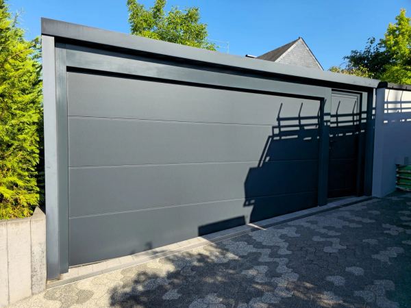 Glattes anthrazitfarbenes Sektionaltor mit Nebentür an einer modernen Flachdachgarage