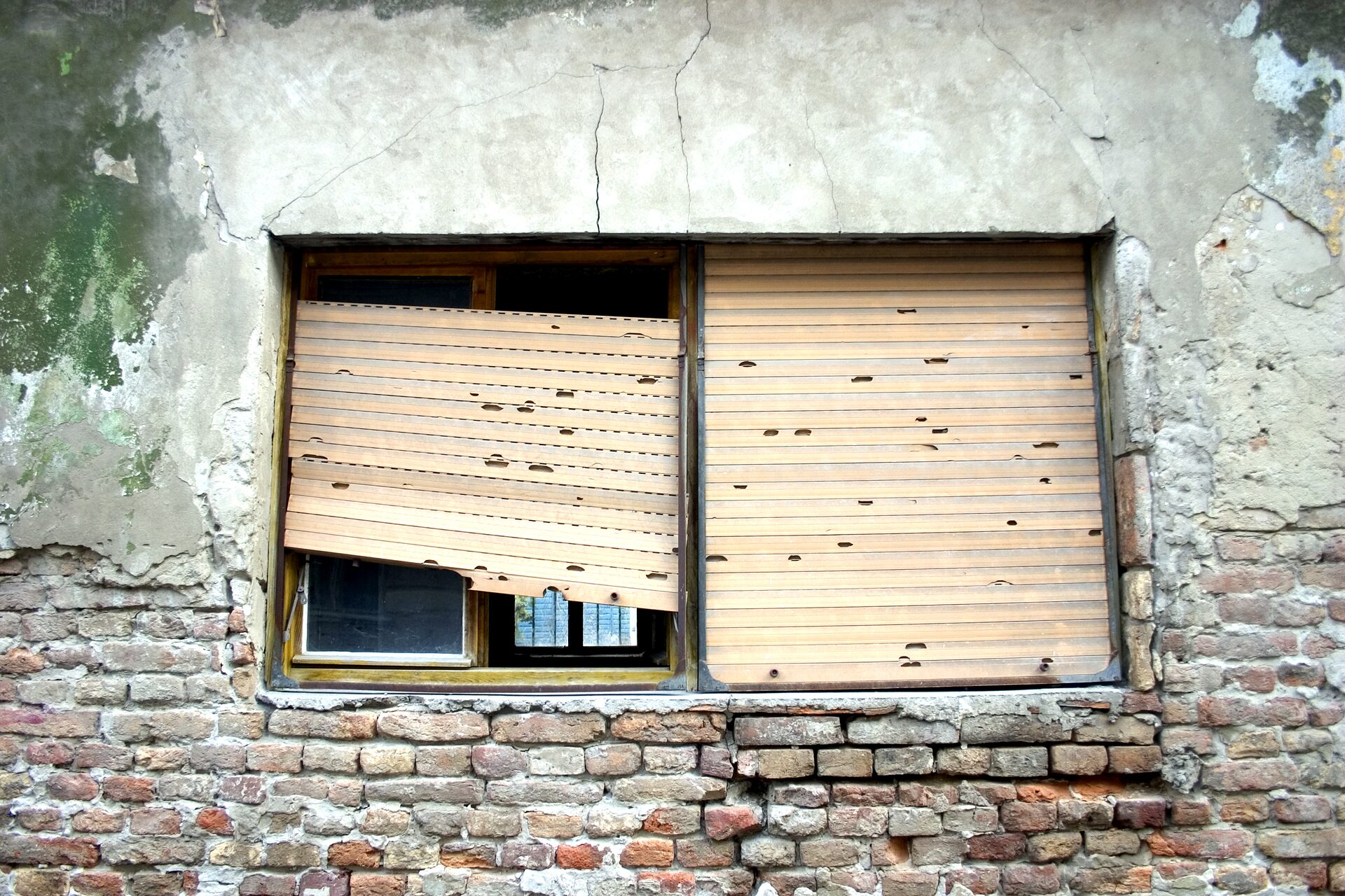 Wooden roller shutter with broken, sagging slats in front of a window on a worn brick façade