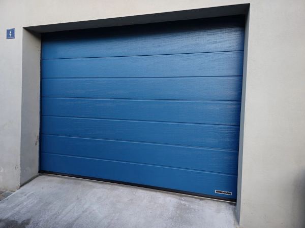 Blaues Hörmann-Sektionaltor mit Sicken und Holzstruktur, graue Putzfassade
