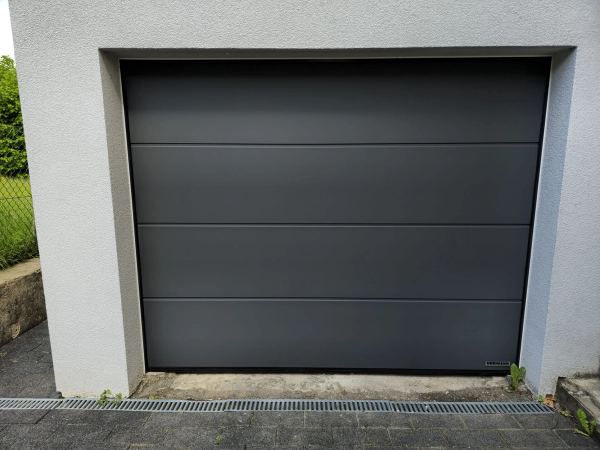Anthrazitgraues Sektionaltor mit vier Paneelen an grau verputzter Fassade, eingezäunter Garten