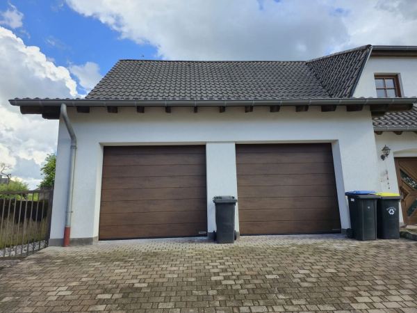 Zwei braune Sektionaltore in Holzoptik an einer weißen Garage mit Ziegeldach