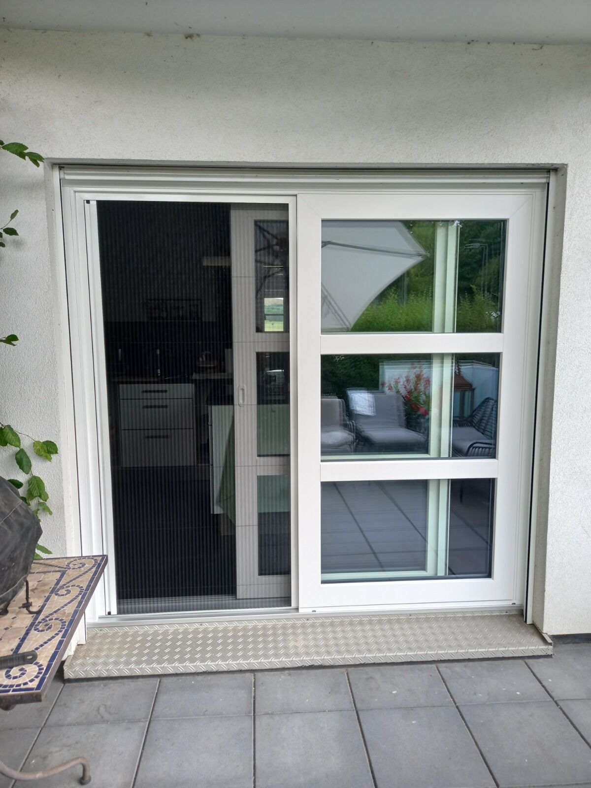 Weiße Schiebetür mit Sprossen, Plissee-Insektenschutz und Riffelblech-Schwelle auf einer Terrasse