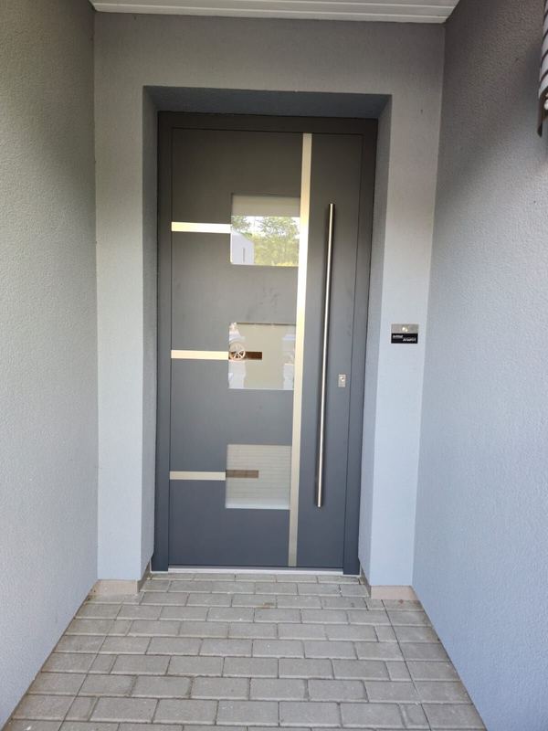 Graue Haustür mit drei kleinen Glasfeldern, Edelstahlband und Lisenen, zurückgesetzter Eingang mit grauem Putz